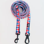 A coiled dog leash with a red, white, and blue gingham pattern, featuring black metal swivel clamps on a plain white background.