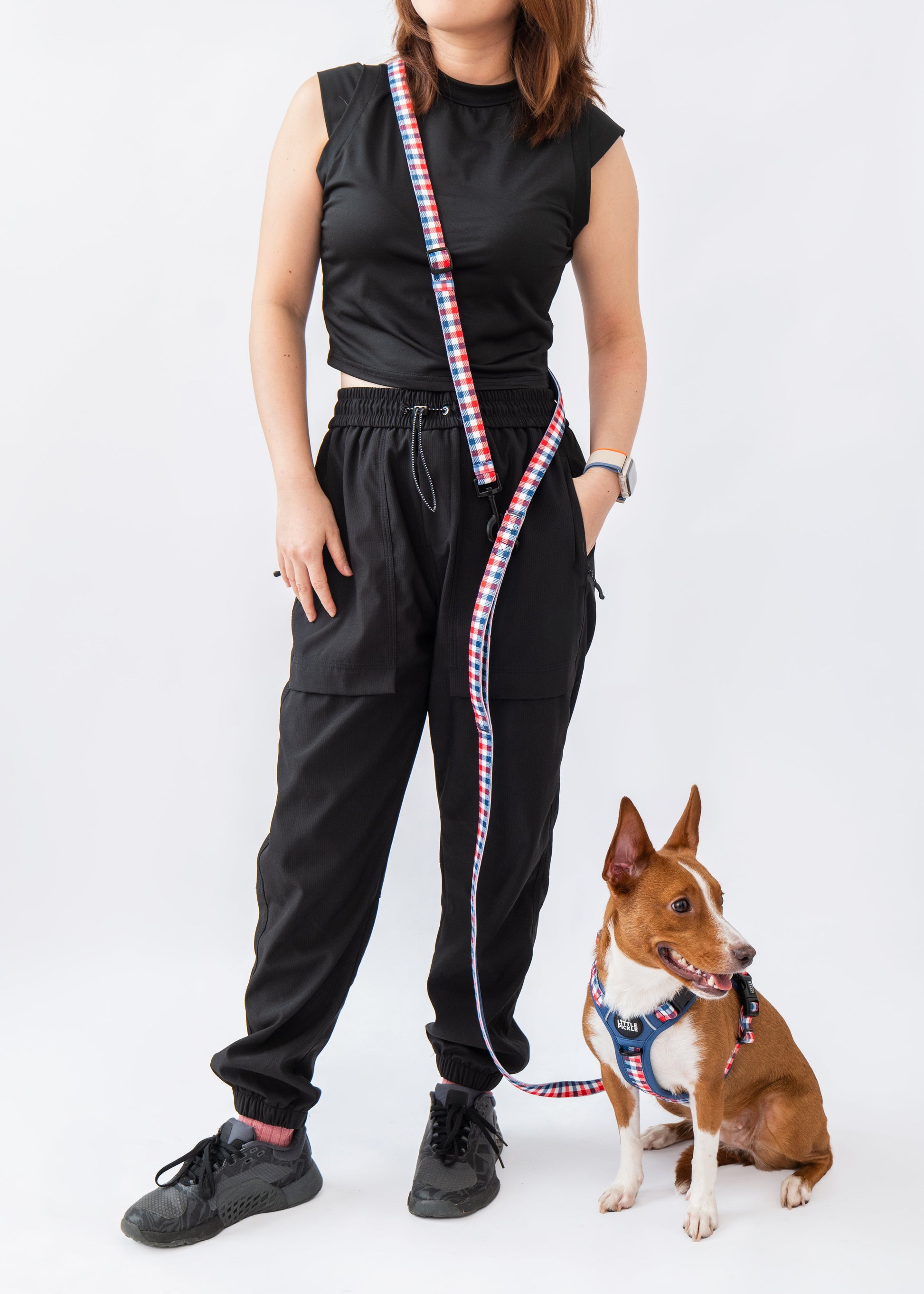 A woman wearing a black sleeveless top and black jogger pants stands next to a small brown and white dog wearing a blue harness, holding a red, white, and blue striped leash, against a plain white background.