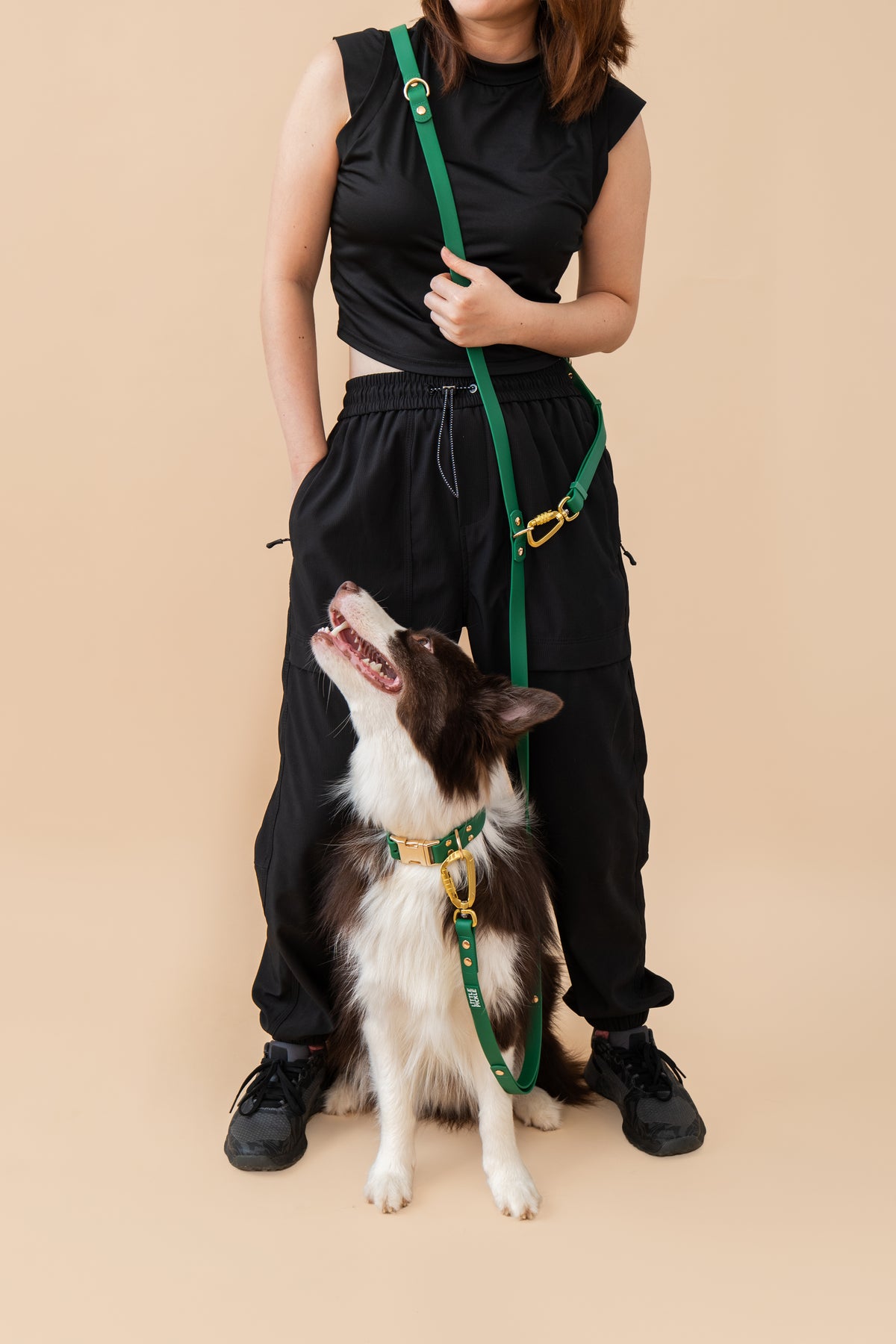 A person wearing a black sleeveless top and black sweatpants stands next to a black and white dog wearing a green and gold harness and leash, against a solid beige background.