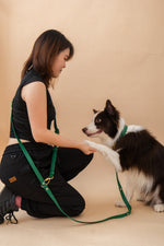 A woman kneels on a beige studio floor, extending her paw to a black and white dog wearing a green leash harness and matching green leash. The woman is dressed in a black sleeveless top and black pants.